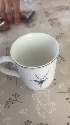 A video shows hot water being poured into a mug. The mug is white with a blue bird and flower design on it. It sits on a patterned tablecloth. The water is clear and appears to be steaming. As the water fills the mug, it mixes with what appears to be instant coffee or tea, creating a light brown beverage. The pouring continues until the mug is nearly full.