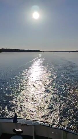 The video shows a bright sunny day with a boat moving forward on the water, leaving a sparkling trail of water behind it. The sun is shining brightly in the sky, reflecting off the water. On the left and right sides of the water, there are small sections of land, with trees and bushes visible. The boat's movement creates a ripple effect on the water, which catches the sunlight and glistens. The sky is clear and blue, and the overall scene depicts a serene and picturesque landscape.