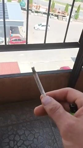 The video shows a close-up of a hand holding a lit marijuana cigarette. The hand is positioned in the foreground, obscuring the view of a balcony railing and a brown wooden railing. In the background, through a metal grate, a parking lot with several cars and buildings can be seen. Further in the distance, a line of trees forms a backdrop under a cloudy sky. The cigarette is held up towards the sky, and the end of it appears to be lit and smoldering. The video is short and shows the cigarette being held steady.