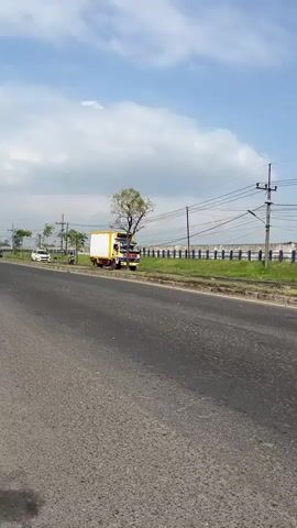A light-colored truck with a white box is parked on the side of a road. The truck has yellow and red accents. A tree with sparse leaves is behind the truck. Further down the road, there are a few white cars and motorcycles. The road is paved, and there is a grassy median with a blue fence separating it from what appears to be a field or industrial area. Power lines and utility poles are visible in the background. The sky is partly cloudy.