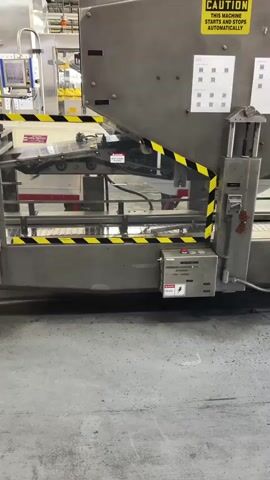 The video shows an industrial packaging machine in operation. The machine is predominantly made of stainless steel and features yellow and black hazard striping. A conveyor belt is visible moving inside the machine. Control panels with buttons and indicator lights are also present. On the top of the machine, a yellow caution sign reads "THIS MACHINE STARTS AND STOPS AUTOMATICALLY." Several QR codes are affixed to the machine's surface. The environment appears to be a factory or warehouse, with a concrete floor and other industrial equipment visible in the background. The video focuses on the mechanical operation of the machine without any human interaction.