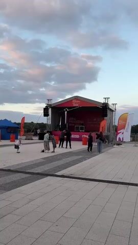 The video shows an outdoor stage setup at what appears to be a festival or event. The stage has a red backdrop with yellow text that reads "ДЕНЬ МОЛОДЁЖИ 2026" (Day of Youth 2026). A DJ is present on stage, operating equipment. Several people are gathered in front of the stage, some watching the DJ, others interacting with each other. One person in the foreground, wearing a white and black patterned outfit, is walking. Another person, wearing a black hoodie, is standing. To the left, a young child in a white dress is standing. There are also flags and event signage visible around the stage area. The sky suggests it is late afternoon or early evening, with some clouds present.