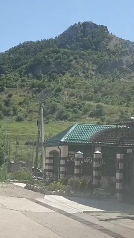 The video shows a static shot of a residential building with a green roof and a decorative fence in front of a mountain. The building has white and black striped pillars and decorative iron gates. There are small evergreen plants planted in front of the fence. A power pole with wires is visible to the left of the house. The mountain behind the house is covered in green vegetation and rocks, and appears to have a cross or monument on its summit. The sky is clear and blue.