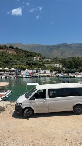 A white minivan is parked on a dirt path next to a body of water filled with boats. The water is calm and a vibrant green color. In the background, there are green hills covered with trees and some buildings scattered amongst them. The sky is clear blue with a few white clouds. The scene appears to be a coastal or lakeside village on a sunny day.