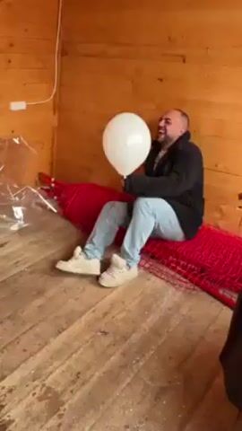 A man with a shaved head and beard, wearing a black jacket and light blue jeans, sits on a red net on a wooden floor against a wooden wall, holding a white balloon and laughing. A plastic sheet is visible to his left.