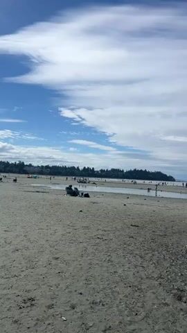 The video shows a wide, sandy beach with scattered puddles of water and some pebbles near the foreground. In the distance, there is a body of water with a distant tree line. The sky is mostly blue with white, wispy clouds. Several people are visible on the beach and in the shallow water, engaged in various activities. Some are walking, others appear to be sitting or playing. There are a few beach chairs and umbrellas scattered around. The overall scene depicts a busy, daytime at a beach.