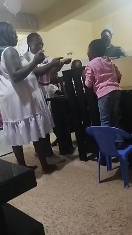 The video shows a casual gathering of people, likely a celebration, in what appears to be a living room. Three adult women are visible, two standing, and one leaning on a dark wooden table. A young child in a pink hoodie and jeans is also leaning against the table, engaging with the adults. A man is partially visible behind the table, seemingly observing. There's a blue plastic chair near the table and balloons can be seen near the ceiling, suggesting a party atmosphere. The room is carpeted and there's a dark-colored coffee table visible in the foreground. The walls are painted a light yellow color. The overall scene seems relaxed and cheerful.