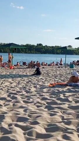 This is a wide shot of a crowded beach on a sunny day. The sand is light-colored and rippled, with a few people lying on towels and beach chairs scattered across it. In the foreground, a person in a white hat and blue swimsuit is lying down, facing away from the camera, with an orange towel beside them. Further back, two people are standing near a green beach umbrella. One person is wearing an orange swimsuit and appears to be walking, while the other, a man, is wearing a striped shirt and shorts and is walking away from the camera.

Beyond the sandy area, there is a body of water, likely a river, with several people swimming or wading in it. The opposite bank of the water is lined with lush green trees, suggesting a natural landscape. Two more green beach umbrellas are visible near the water's edge. Many people are gathered on the beach, some sitting, some standing, and some walking. The overall atmosphere appears relaxed and leisurely, typical of a summer beach day. The sky is a clear, bright blue with a few wispy white clouds, indicating fair weather. The sunlight casts shadows on the sand, suggesting it is likely late morning or early afternoon. The presence of trees along the riverbank and the general style of the umbrellas and beachgoers are consistent with a European setting, specifically Kremenchuk, Ukraine, as indicated by the context. There is no visible text in the media file.