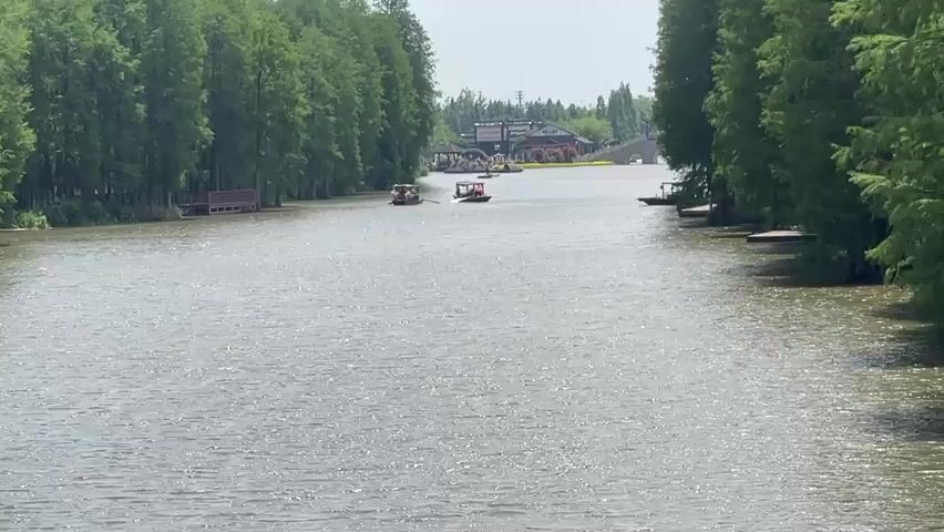 The video shows a wide, calm river with trees lining both banks. There are several small boats moving slowly in the water. In the distance, there's a building complex and a bridge over the river. The sky is overcast.