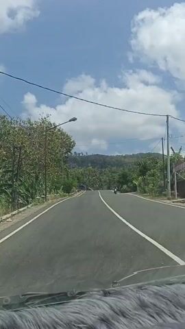 The video shows a first-person perspective from inside a vehicle, looking out at a paved road. The road is bordered by trees and vegetation. There is a street light on the left side of the road and power lines stretching across the sky. In the distance, there is a hill covered in trees. A motorcycle is visible on the right side of the road, traveling in the same direction as the vehicle. The sky is blue with scattered white clouds. The dashboard of the vehicle is visible at the bottom of the frame, covered with a gray furry material.