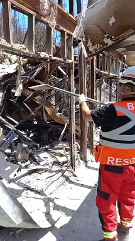 A rescue worker wearing a white helmet, red pants, and an orange vest with the word "RESCATE" on the back is clearing debris from a fire-damaged structure with a metal bar. The structure's frame is visible, with the interior gutted and filled with charred remains. Another worker stands in the background, also dressed in protective gear. The environment shows signs of extensive fire damage.