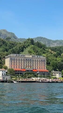 A body of water with ripples and waves is in the foreground. A large, multi-story building with a tan facade and many windows sits on the shore in the middle ground. The building is situated on a hillside covered in green trees. A bright blue sky is visible above the building and trees. Several smaller buildings and white umbrellas are also visible along the shore in front of the large building. Boats are docked near the shore. The scene appears to be a sunny day.