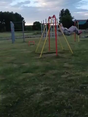 A person in a light-colored tracksuit is swinging on a swing set in a park. The swing set has red and yellow metal poles. The park has a grassy ground with scattered patches of dirt. In the background, there are trees, a fence, a sandbox, and other playground equipment, including a pull-up bar and what appears to be outdoor exercise machines under a blue awning. The sky is cloudy, suggesting it is either early morning or late afternoon. The overall mood is casual and playful. There is no visible text in the video.