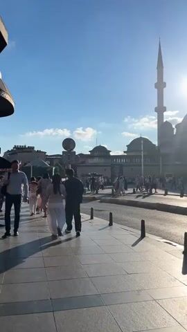 The video pans across a street scene with a large mosque in the background. The sun is bright and positioned behind the mosque, creating a lens flare effect. Several people are walking on the sidewalk and crossing the street in front of the mosque. A red tram is visible to the right of the mosque. The architecture of the mosque includes domes and minarets. The sky is blue with some clouds. The foreground shows a tiled sidewalk and a street with a curb.