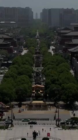 From an elevated perspective, a straight avenue stretches into the distance, framed by symmetrical rows of trees, classical red-roofed buildings, and modern high-rise buildings on the horizon. The avenue features various statues and decorative structures along its length. People stroll along the walkway and cross the street in the foreground. Cars are also visible on the roads. The weather appears hazy or overcast.