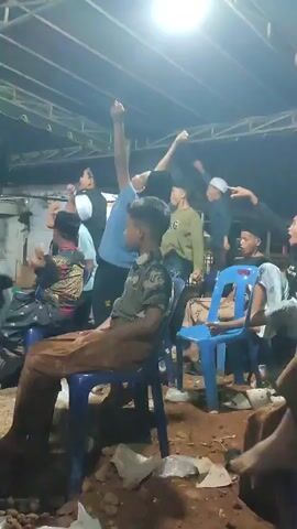 The video shows a group of men, mostly young, gathered under a covered structure, possibly an outdoor event or gathering. The lighting suggests it is nighttime, with a bright ceiling light illuminating the scene. Most of the men are seated on blue plastic chairs, some appearing to be engaged in a performance or activity, with raised hands and enthusiastic gestures. The ground is uneven and appears to be dirt with some scattered debris. The men are dressed in casual attire, with some wearing skullcaps, typical of religious or cultural gatherings. One young man in the foreground is wearing a brown uniform-like shirt. The atmosphere seems lively and celebratory.