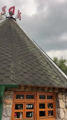 This media file is a vertical video clip capturing the exterior of a rustic establishment in Mekića Kula, Montenegro. The scene showcases unique, conical-roofed buildings constructed with rough-hewn stone walls and dark, shingled roofs. One building features a prominent wooden window with multiple panes, adorned with what appear to be decorative cutouts of cat faces. Above this building, a red neon sign with Cyrillic letters spelling "КАФЕ" is visible.

The setting suggests an outdoor seating area, with wooden picnic tables and benches arranged on a stone-paved ground. A large, beige umbrella is folded and stands to the right of the frame, partially obscuring the view of the surrounding greenery. Beyond the buildings, a dense forest of evergreen trees, including fir trees, dominates the background. A white car is parked in the distance, and a wooden fence winds its way through the landscape.

The weather appears overcast, with a cloudy sky dominating the upper portion of the video. The overall mood is peaceful and serene, evoking a sense of being in a secluded, natural environment. The time of day is difficult to ascertain precisely due to the cloudy conditions, but it seems to be daytime. There are no people visible in the clip.