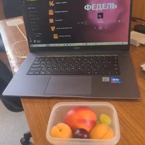 The video is a close-up shot of a laptop on a wooden table. In front of the laptop is a clear plastic container filled with fruit, including apricots, a plum, a peach, and a grape. The laptop screen displays an application with a playlist titled "FEDEL". The laptop is open and appears to be in use. The lighting suggests an indoor setting, possibly an office or study.