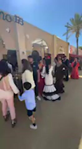 The video captures an outdoor scene during daylight hours, under a bright, sunny sky. The setting appears to be a paved area or street in front of buildings with light-colored walls and arched doorways. A palm tree is visible in the distance.

The primary subjects are a group of people, predominantly women and girls, engaged in flamenco dancing. They are dressed in traditional, elaborate flamenco attire, including ruffled dresses in colors such as white with black trim, red, and brown. Some of the dancers also wear wide-brimmed hats adorned with flowers. They perform rhythmic arm and body movements characteristic of flamenco. Briefly at the beginning, a man in a black t-shirt and black pants walks past, gesturing with his hand. A young boy in a light blue shirt and dark shorts is also partially visible in the foreground at times.

The activity taking place is a cultural performance or gathering centered around flamenco dance. The overall mood is festive and lively, reflecting the traditional art form. On a building in the background, the letters "BB" are partially visible. The flamenco attire and architecture are strong indicators of a Spanish or Andalusian location, consistent with Jerez de la Frontera, Spain.