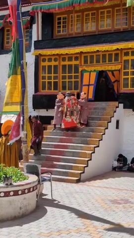 Three people wearing masks stand on stairs. Two of the people, one on each side, wear brown robes and have face masks. The person in the middle wears a red and white robe. The person on the left appears to be holding a long stick. The stairs have red, yellow, and white stripes. In the background, there is a building with yellow-trimmed windows and colorful flags. To the left, there is a potted plant and a chair. To the right, several people are sitting on the ground. The ground is paved with interlocking bricks.