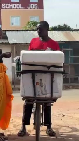 The video shows a man on a bicycle loaded with two white styrofoam coolers and boxes strapped to the rack over the rear wheel. The man is wearing a red t-shirt and dark pants and sandals. A woman wearing an orange wrap-around skirt stands beside him, facing him and holding what looks like money or a receipt. The background includes buildings, one of which is labeled "SCHOOL, JOS." The street is unpaved.