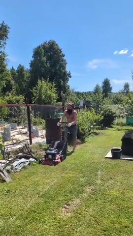 A shirtless man with a beard and a bandana on his head is pushing a red and black lawnmower across a grassy yard. He is wearing dark shorts and sandals. The lawnmower is in motion, and the man appears to be actively mowing the grass. The background consists of trees, bushes, a fence, and some gardening equipment. The sky is blue with some white clouds. The scene suggests a sunny day. The video is very short, lasting only a few seconds, and captures a brief moment of yard work.