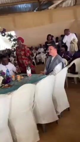 The video captures a scene inside what appears to be a banquet hall or event space, where a group of people is gathered. The hall has white chairs and tables. The table in the foreground is decorated with flowers. A man wearing glasses, a white shirt, and a gray robe-like garment is seated at the table, smiling slightly as he looks to his right. Around him, other people are standing or seated. A woman in a colorful patterned dress is serving food from a tray to the man. Two men in the background stand and watch. The overall atmosphere seems pleasant and celebratory.