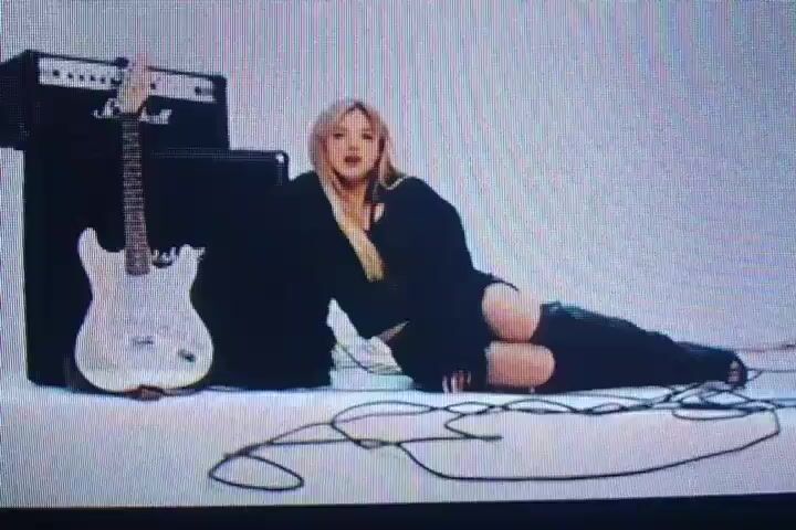 The video is comprised of four short clips. The first clip shows a woman with blonde hair lying on the floor next to a guitar and a large amplifier. She is wearing a black outfit with ripped pants. The second and third clips show the same woman in pajamas, speaking and gesturing with her hands. A man in pajamas and glasses is visible in the background of the second and third clips, holding a plate of donuts. The fourth clip shows the woman in pajamas, speaking to another person who is off-screen. The environment in the second, third, and fourth clips appears to be a room with pink walls.