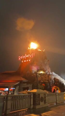 The video depicts the Rainforest Cafe with a simulated volcano erupting in flames. The restaurant’s name is illuminated in red against the dark sky. A white picket fence lines a walkway in front of the building. Two people wearing yellow raincoats walk along the walkway. The volcano structure has multiple openings where flames shoot out. Smoke rises into the night sky. A street lamp provides additional light to the scene.