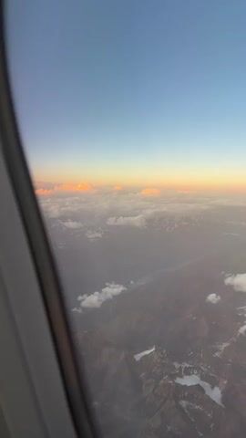 The video shows an aerial view from an airplane window. The camera is positioned at an angle, looking out at a landscape covered in clouds and mountains. The sky transitions from a deep blue at the top to a pale orange and yellow near the horizon, indicating either sunrise or sunset. Scattered clouds are visible below, some illuminated by the sun with a soft orange glow, while others appear white and fluffy. The mountainous terrain beneath the clouds is partially obscured, but glimpses of rocky surfaces and snow-capped peaks can be seen. The edge of the airplane window frame is visible on the left side of the video, suggesting the perspective of a passenger. There are no people or specific actions captured in the frame; the focus is on the natural scenery from a high altitude.