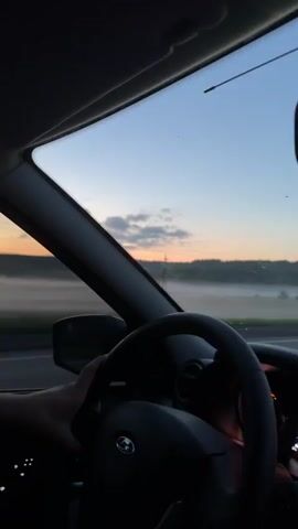 This video shows a first-person view from the inside of a car as it travels along a highway. The driver's hands are visible on the steering wheel, and the passenger's arm is resting on the door. Outside the car, a misty landscape unfolds under a soft, dusky sky. The sun appears to be setting or rising, casting a warm orange glow on the horizon. Scattered clouds drift across the pale blue sky. The mist hangs low over fields and trees, creating a serene and slightly mysterious atmosphere. In the distance, there are power lines and poles, suggesting a rural or semi-rural setting. The overall mood is peaceful and contemplative, likely evoking a sense of journey or quiet reflection during the early morning or late evening. The location is identified as Minderevo, Russia. There is no visible text in the video.