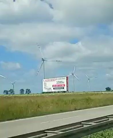 This is a video taken from a moving vehicle, likely a car, showing a landscape with several wind turbines. The foreground is dominated by a road and a green barrier. In the background, there is a field of tall grass and some trees, with the wind turbines scattered across the horizon. The sky is partly cloudy, with some patches of blue. A large billboard is visible in the distance, advertising mattresses. The video captures the movement of the vehicle, with the scenery passing by. The wind turbines are tall, white structures with rotating blades, characteristic of wind farms. The green barrier in the foreground appears to be a noise barrier or safety fence for the road. The overall impression is of a rural or semi-rural setting with a focus on renewable energy.