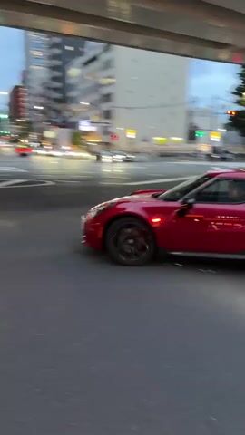 A red Alfa Romeo 4C sports car drives away from the camera on a city street in Minato, Japan, during dusk or early evening.

At 0:00, the car is seen in profile, driving from left to right under a concrete overpass. The car has black wheels and a black lower body trim. The background shows city buildings with some lights on.

From 0:01, the car is viewed from the rear as it continues under the overpass and turns right onto another street. Two orange and white striped bollards are visible on the roadside in the foreground. Other cars and a black van are seen in the background on the street, with buildings and trees lining the area.

At 0:03, a black van with a distinctive yellow/lime green stripe and some unreadable white text on its side passes close to the camera, causing a motion blur effect. The text appears to be "HOTEL" mirrored. The scene is set in an urban environment with multi-story buildings and an overhead concrete structure.