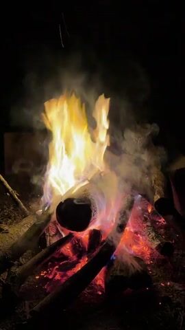 The video captures a campfire burning in the dark. There are several logs arranged in a pile, with flames rising high, interspersed with smoke. The fire radiates a warm, red glow at the base and transitions to bright yellow flames higher up. A sound of laughter is heard in the background.