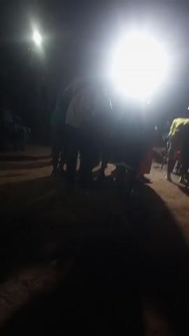 The video shows an outdoor gathering at night. The scene is dimly lit with strong spotlights. Several people are standing and interacting in the foreground. Further back, there are people seated in chairs, some possibly watching or listening to a DJ or musician standing behind a setup that could be sound equipment. The ground appears to be dirt or sand. The overall atmosphere suggests a social event with music, though the specific details are unclear due to the lighting conditions.