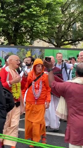The video shows a religious procession or parade with several people participating. In the foreground, a bald man wearing glasses and a red jacket is speaking into a microphone. He is adorned with yellow flower garlands around his neck. To his right, another person in an orange hooded sweatshirt and orange skirt also wears similar flower garlands. Behind them, other individuals are visible, some holding musical instruments, including what appears to be a drum. Further back, a decorated chariot with figures on it is being pulled, and a crowd of onlookers lines the path. The environment appears to be an outdoor urban setting with trees and buildings in the background. The overall atmosphere suggests a festive or devotional gathering.