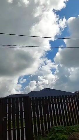 The video shows a view of the sky with clouds and some buildings. In the foreground, there is a wooden fence. Behind the fence, there are roofs of houses and a power pole with wires. The sky is blue with white and gray clouds. The sun is visible through the clouds, creating highlights. The overall scene suggests an outdoor environment, possibly a residential area, with varying weather conditions.