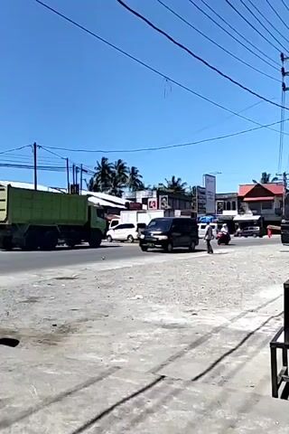 The video shows a daytime street scene under a clear blue sky. A large green dump truck is parked on the left side of the road, and a black van is driving towards the right. A person in a white shirt is crossing the street. Two individuals on a red scooter pass by on the left side of the road. Utility poles and power lines are visible overhead.  Palm trees and buildings line the opposite side of the street. The road surface appears uneven and cracked. The video seems to be taken from a slightly elevated position, possibly from a sidewalk or building.