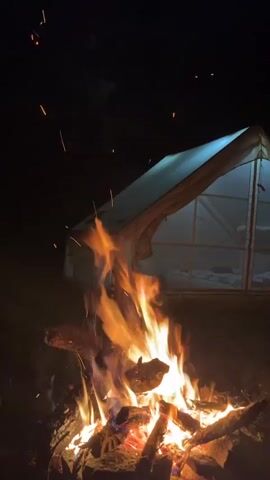 A nighttime scene depicts a campfire in the foreground with a canvas tent illuminated from within in the background. The campfire is ablaze with bright orange and yellow flames, casting a warm glow on the surrounding logs and ground. Sparks from the fire ascend into the dark sky, creating small, glowing streaks.

The tent, a triangular structure, is a pale color, possibly white or light beige, and its large windows are lit, suggesting occupancy. Through the windows, glimpses of what appear to be sleeping bags or beds can be seen. The tent is set against a dark background, indicating it is nighttime. The overall setting appears to be an outdoor campsite.

The time of day is clearly night, with a dark sky and artificial light from the tent. There are no visible weather cues, but the presence of a campfire suggests cooler temperatures or a desire for warmth and ambiance. The location is identified as Simeonovets, Bulgaria. The scene evokes feelings of coziness, relaxation, and perhaps adventure associated with camping. No text is visible in the media.