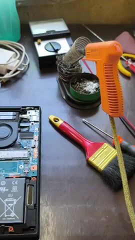 This media file shows an open laptop on a wooden surface. The laptop's internal components are exposed, including the motherboard, fans, battery, and what appears to be a solid-state drive.

**Objects:**
*   **Laptop:** The main subject, with its bottom casing removed, revealing the intricate internal electronics. The battery is labeled "Li-ion 00" and has a capacity of "min. 5080mAh." There's also a "Lenovo" logo visible on the battery.
*   **Soldering Iron:** An orange soldering iron with a coiled wire for its stand is prominent.
*   **Soldering Tin:** A can of soldering tin is next to the soldering iron.
*   **Paintbrush:** A red-handled paintbrush with a gold ferrule and black bristles is also on the surface.
*   **Screwdriver:** A screwdriver is visible near the paintbrush.
*   **Multimeter:** A digital multimeter is in the background.
*   **Power Adapter:** A white power adapter with cables is in the background.
*   **Green Object:** A green object, possibly a bottle or container, is in the upper left background.

**Activity/Event:**
The presence of a soldering iron and tin suggests that some repair or modification work is being done on the laptop. The tools laid out indicate an electronic repair or maintenance scenario. The laptop is disassembled, allowing access to its internal components for potential soldering, cleaning, or component replacement.

**Notable Details:**
*   **Lighting:** The lighting appears to be artificial, suggesting an indoor setting.
*   **Setting:** The wooden surface implies a workbench or desk.
*   **No People Visible:** The media focuses solely on the objects and the laptop's internals.
*   **Text Visible:**
    *   "Li-ion 00" and "min. 5080mAh" on the battery.
    *   "Lenovo" logo on the battery.
    *   Various labels and certifications on the motherboard and battery, including "CE" and an EU symbol.
    *   Text on the soldering iron: "DEKO / HP-3570 / 220-240V~ 50/60Hz 60W".
    *   Text on the multimeter indicating voltage and other measurements.
    *   Text on the power adapter: "120W".

**Location Context:** Tungkob, Indonesia. This information is provided separately and is not visually evident in the media itself.