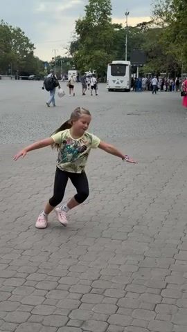 A young girl with blonde hair in a ponytail, wearing a yellow t-shirt and black leggings, performs a cartwheel on a paved outdoor area. She jumps and spins, landing briefly on her hands before completing the movement and returning to a standing position. Other people, including adults and children, are visible in the background walking and standing near buses and trees. The ground is made of interlocking gray pavers. The lighting suggests it is daytime.
