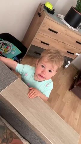 The video shows a young child with blonde hair, wearing a light blue t-shirt with pink flower designs, standing and reaching over a wooden surface. The child's expression is one of surprise or slight distress. In the background, a black toy bin is visible, containing various toys and a remote control. A wooden dresser with black handles is also in the background, along with a beige wooden floor. The child's left arm is resting on a grey textured surface, possibly a couch or chair. The camera angle is from above and looking down at the child.