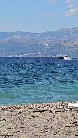 A boat speeds across the ocean, leaving a white wake behind it. The water is a deep blue, with some lighter turquoise closer to the shore. The shore itself is made up of pebbles and rocks, with some larger stones in the foreground. In the distance, there are mountains with some buildings visible on their slopes, and the sky is a clear blue with a few wispy clouds. The overall scene is one of a sunny day at the beach.