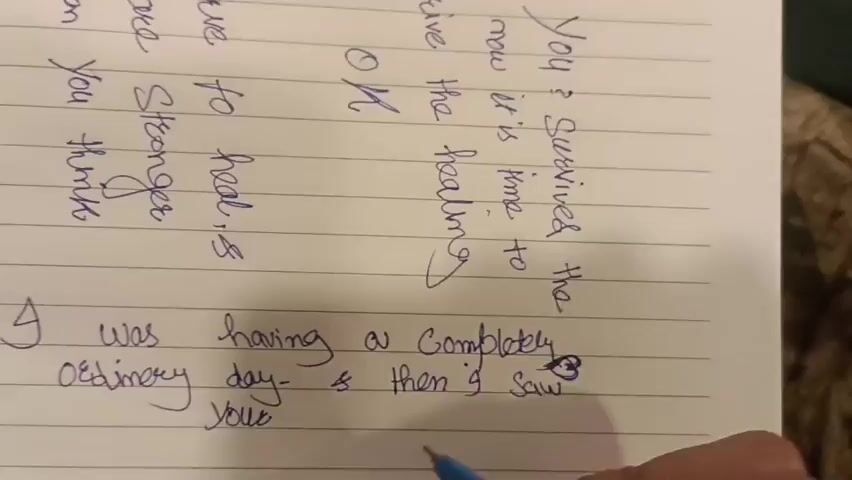The video shows a person's hand writing on a lined notebook. The writing is in blue ink. The notebook page has a watermark. The writing is in cursive and some of the words are difficult to decipher. There are two distinct sections of writing on the page. The top section appears to be a poem or a message, with phrases like "You survived the now it is time to receive the healing" and "are stronger to heal, and than You think". The bottom section is a narrative: "I was having a completely ordinary day - & then I saw your Smile". A portion of a person's arm and hand are visible, holding a blue pen, which is actively writing the last word "Smile". The lighting is dim, suggesting an indoor setting.
