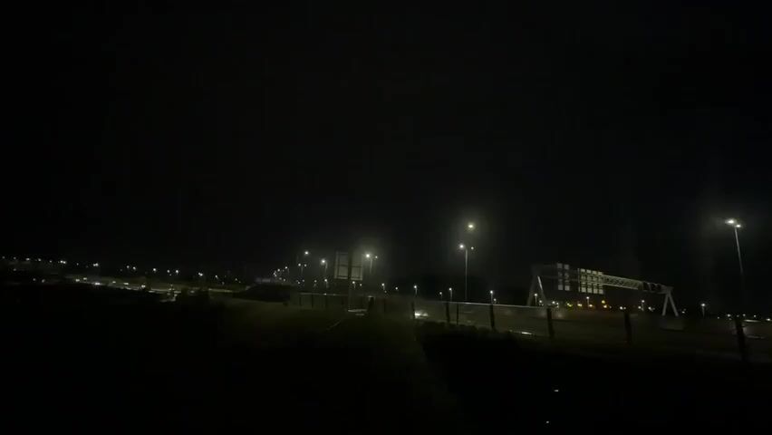 The video shows an outdoor scene at night. The sky is dark, indicating it is nighttime. Several streetlights are illuminated, casting pools of light on the ground and illuminating parts of the surroundings. In the background, there appears to be a road or highway with more lights visible in the distance, suggesting an urban or suburban area. There is also a large, modern structure on the right side of the frame that looks like an overhead road sign or gantry, also illuminated by lights. The foreground is dark, with some undulated ground visible. No people or vehicles are clearly identifiable in the video. The overall impression is of a quiet, possibly deserted, road at night.