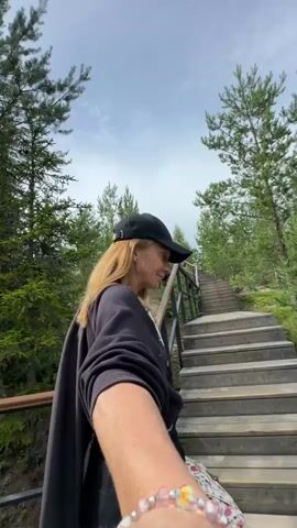 A woman with blonde hair, wearing a black baseball cap and a black sweatshirt, is walking up a wooden staircase outdoors. She is wearing a bracelet on her left wrist. The staircase is surrounded by pine trees and greenery. The sky is cloudy. The video appears to be shot from a first-person perspective.