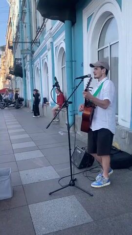 A street performer plays an acoustic guitar and sings into a microphone on a paved sidewalk. He is wearing a white shirt, dark shorts, and a baseball cap. A small amplifier and a trash bin are visible nearby. Several people are walking and standing along the sidewalk, some of whom are observing the performance. Behind the performer, a blue building with arched windows lines the street. The sky above is clear and blue, suggesting daytime. The scene appears to be in an urban environment with historical architecture.