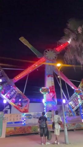 This media file captures a nighttime scene at a fairground or amusement park, likely in Canet de Mar, Spain, given the context. The central focus is a large amusement ride, a spinning pendulum-like attraction with bright red LED lights along its arms and colorful lights on its central tower. Two young people, a male and a female, stand facing the ride, appearing to observe it or wait for it to begin. They are casually dressed, with the male wearing a backpack and the female carrying a tote bag.

The setting is a vibrant, lively atmosphere typical of a nighttime amusement park. The sky is dark, indicating it is after sunset. Palm trees are visible in the background, adding to the tropical or Mediterranean feel of the location. The ground in front of the ride is illuminated by the colorful lights of the attraction, creating a festive and energetic mood.

There is some visible text on a sign at the base of the ride. Though partially obscured and not fully legible, it appears to be instructions or rules related to the ride, with pictograms indicating safety guidelines. The overall impression is one of youthful excitement and anticipation, common to fairground experiences. The focus is on the visual spectacle of the illuminated ride and the people experiencing it.