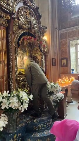 The video shows a man in a green jacket and dark pants bending over to kiss an icon inside a church. He is standing on a patterned rug in front of a large, ornate golden iconostasis. To his left are vases of white lilies. In the background, a row of lit candles is visible, and another person in military attire is kneeling. The church interior features ornate decorations and religious imagery.