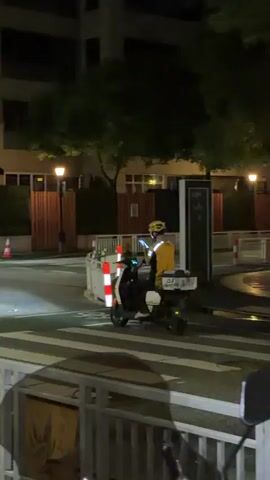 A person on a scooter, wearing a yellow helmet and an orange and white jacket, is stopped at a crosswalk. The person is looking at their phone, which they are holding in their right hand. The scooter has a large storage box on the back. The scene appears to be a street at night, with buildings and trees visible in the background. There are streetlights and the headlights of the scooter are on. The crosswalk is marked with white stripes. There are also traffic cones and reflective bollards visible on the side of the road. A shadowy figure is blurred in motion in the background. The text "GY" is visible on the storage box of the scooter. The overall mood is one of nighttime urban transit.